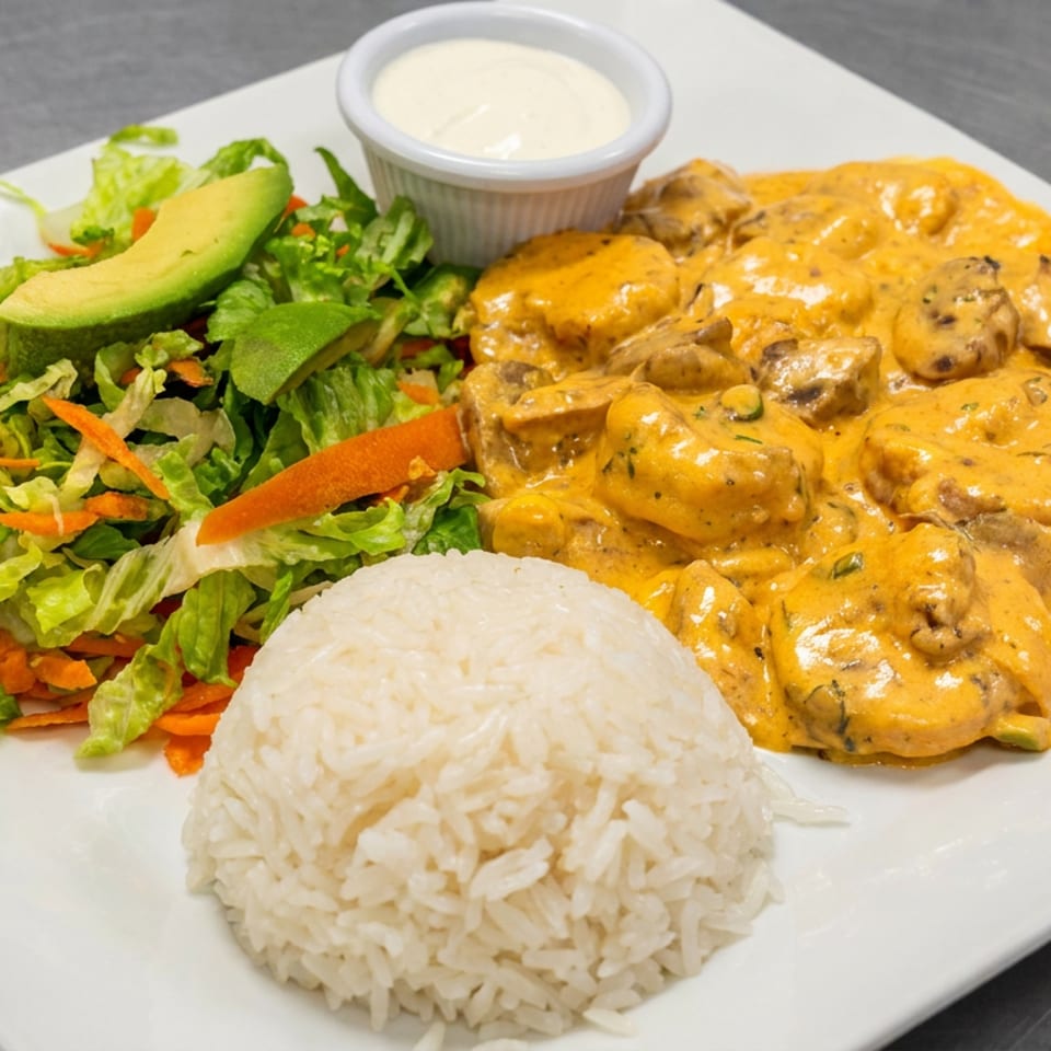 White plate with rice, mixed green salad topped with avocado, and creamy orange mushroom chicken with dipping sauce.